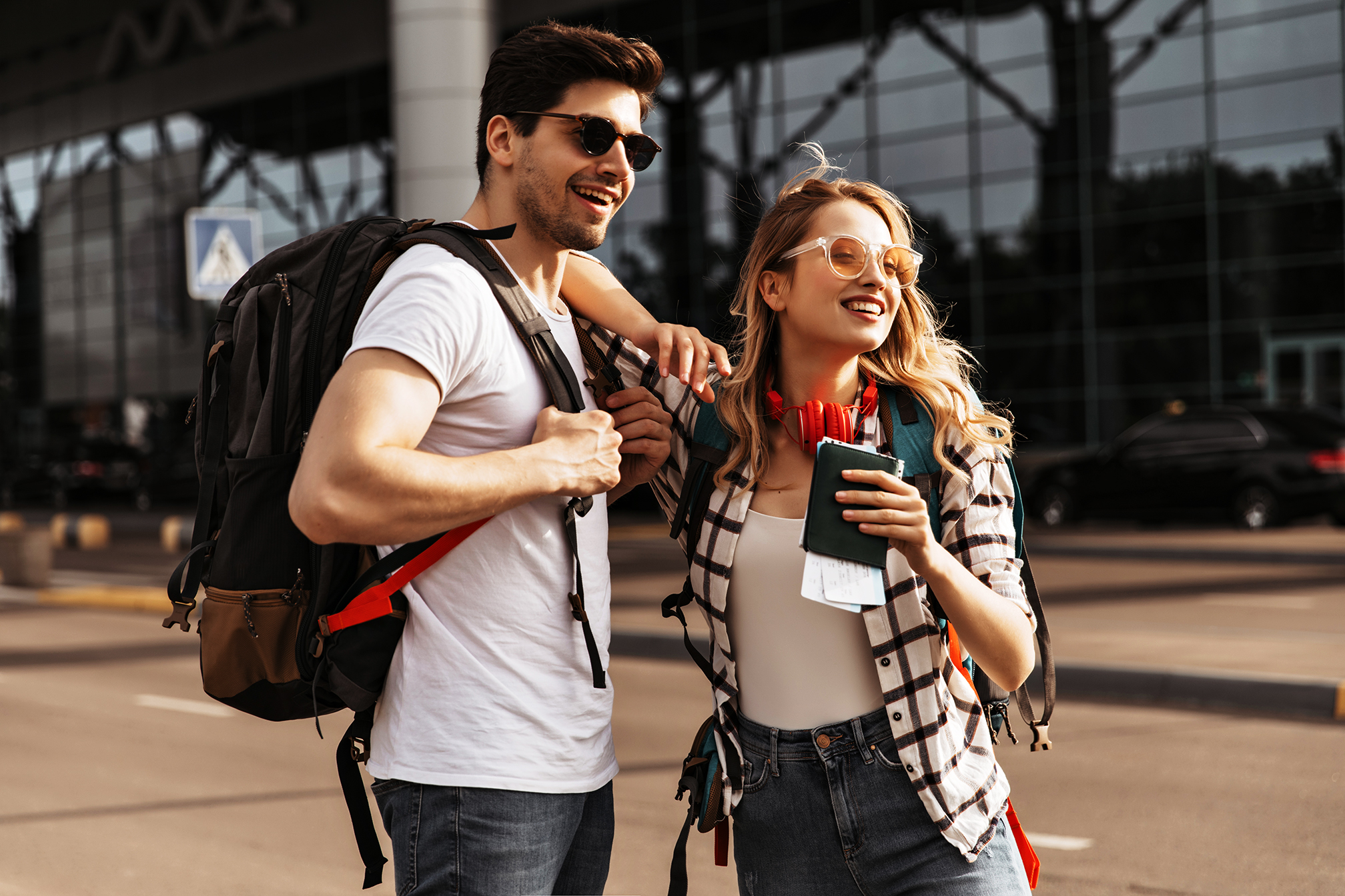 Travelers with backpacks poses near modern airport. Happy blonde woman in plaid shirt, sunglasses holds passport, brunette man in white tee smiles.